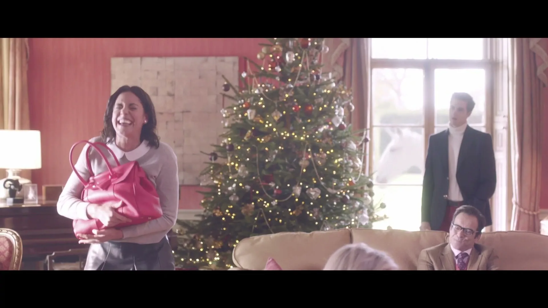 A woman in a grey sweater and white collar holds a red handbag, laughing joyfully in a festive living room with a Christmas tree.