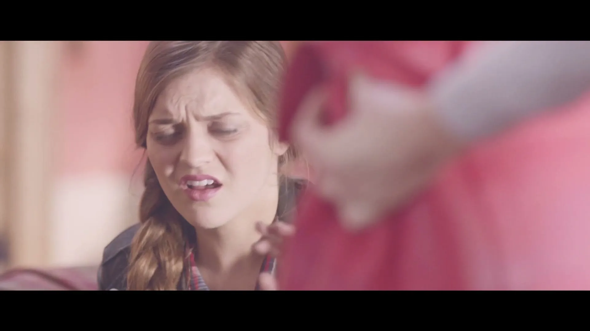 A young woman with a braid and a leather jacket looks disgusted while another person in red is out of focus in the foreground.