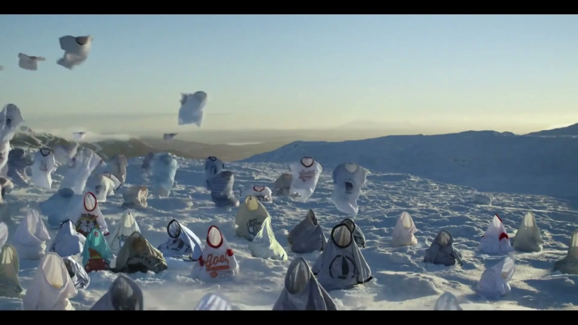 Hundreds of t-shirts, some filled with air, some draped over objects, are scattered across a vast, snowy mountain landscape under a clear sky.