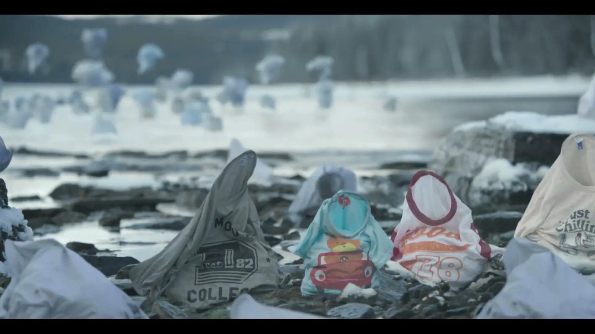 Various t-shirts, some with designs, are arranged on a rocky, snowy shore next to a body of water, resembling small figures.
