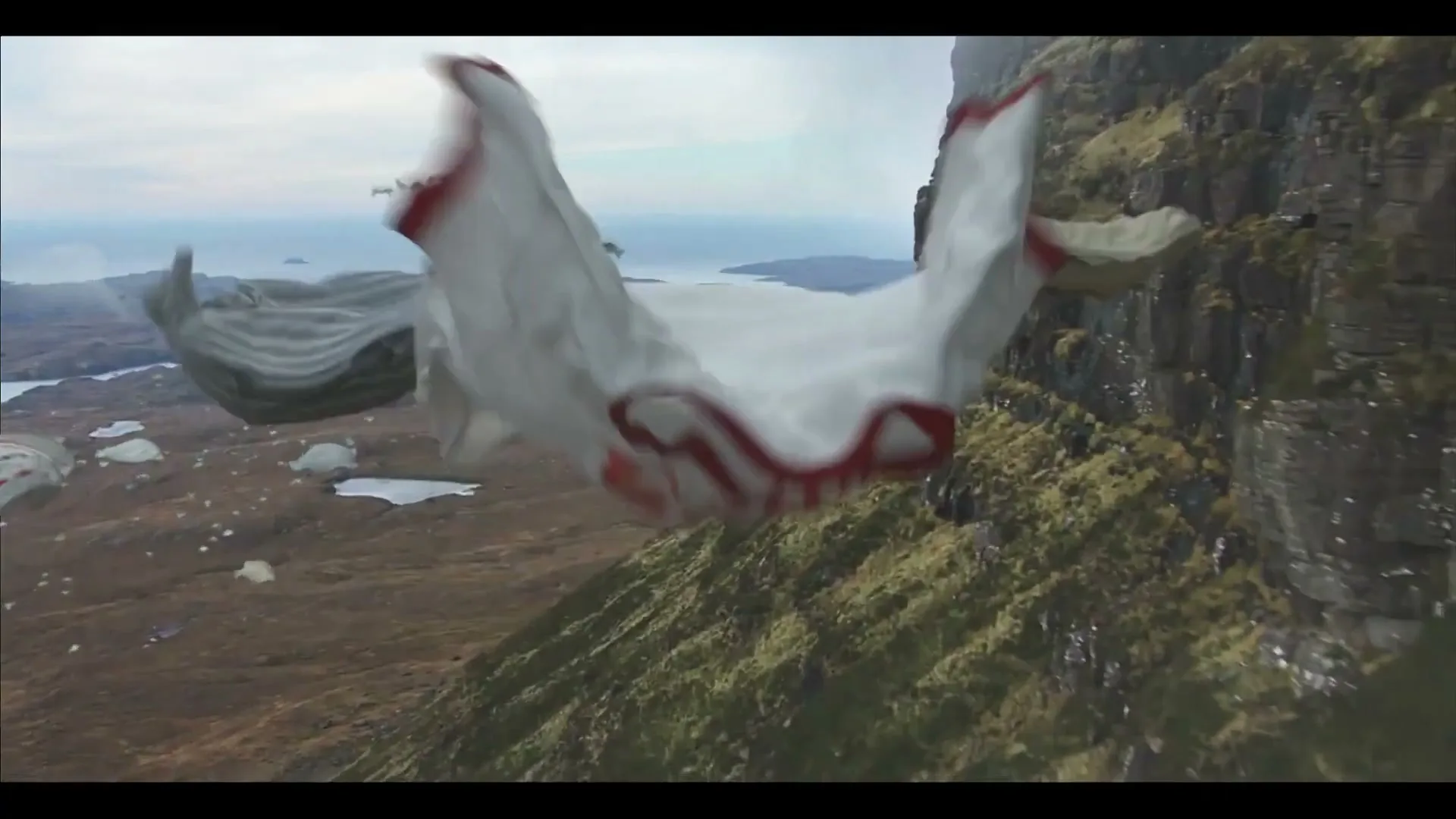 A white t-shirt with red trim flies through the air, resembling a wingsuit, with other t-shirts in the background over a mountainous landscape.