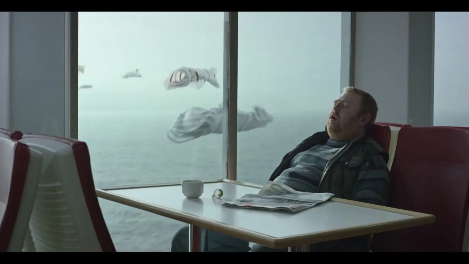A man with a beard sleeps at a table on a ferry, while t-shirts shaped like fish swim past the window in the background.