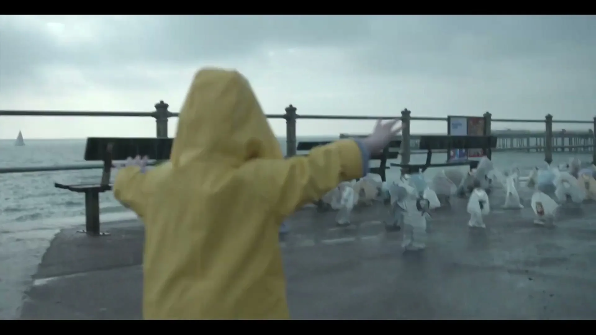 A child in a yellow raincoat runs towards the ocean on a pier, with many white t-shirts on the ground, on a rainy day.