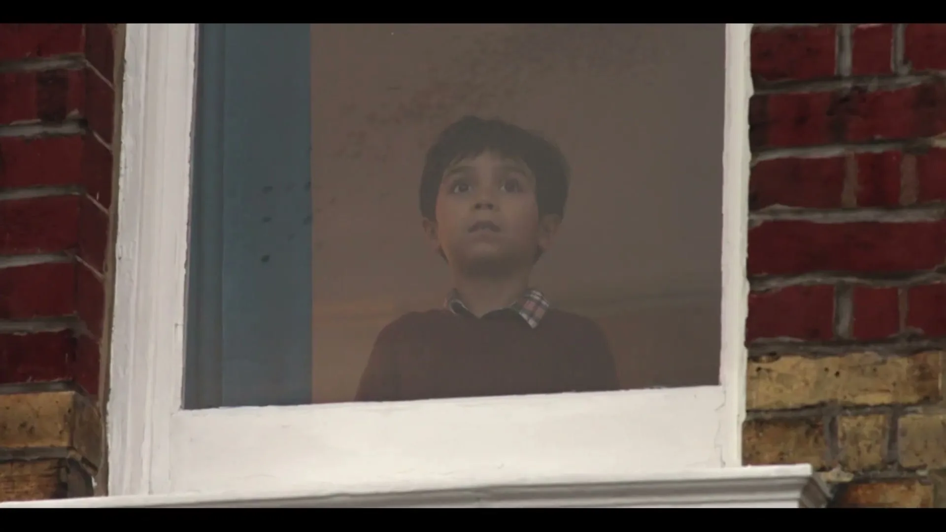 A young boy with dark hair looks up with wide eyes through a window, framed by a brick wall.
