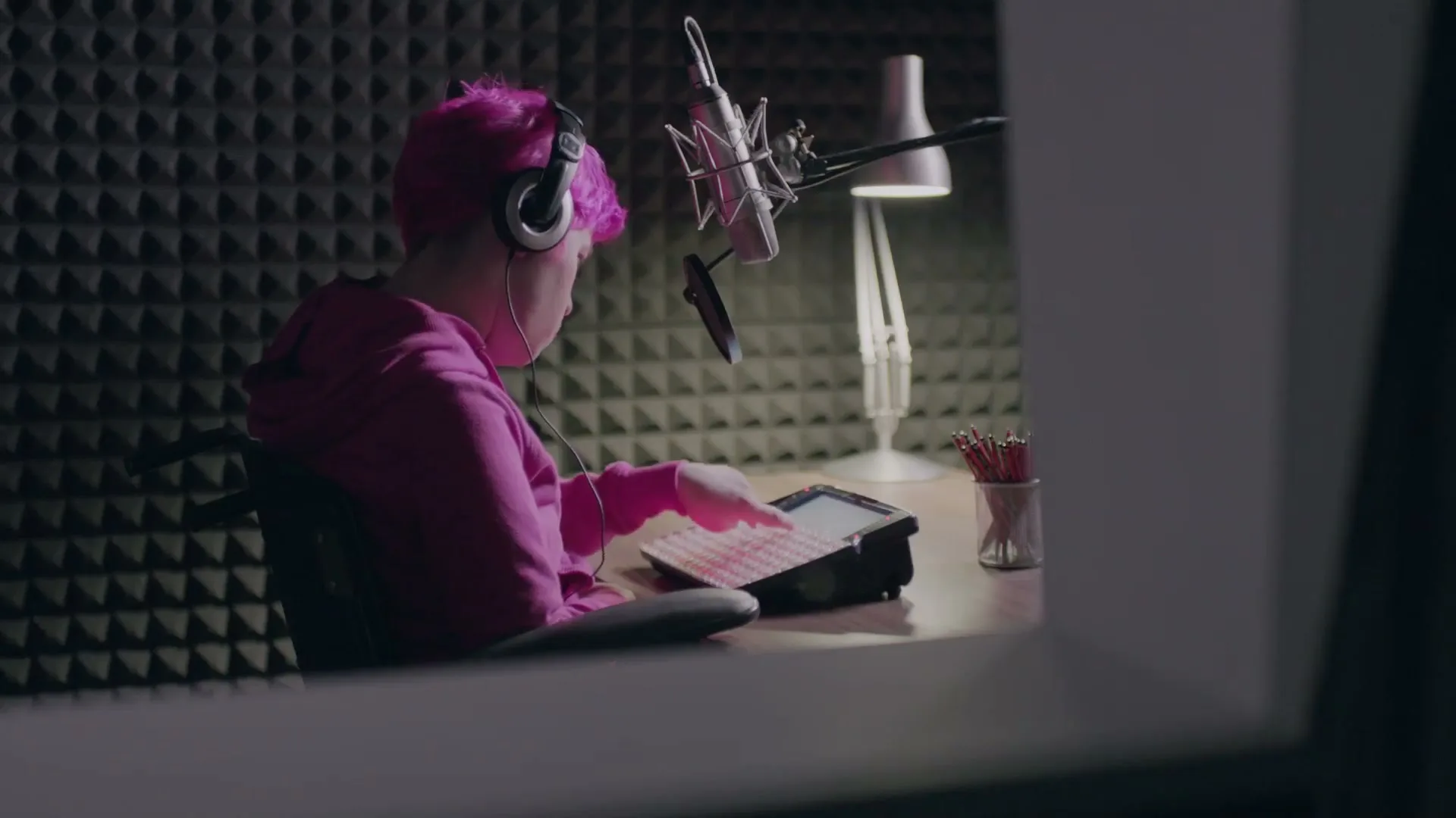 A person with pink hair and headphones sits at a desk in a sound booth, using a communication device with a microphone in front of them.