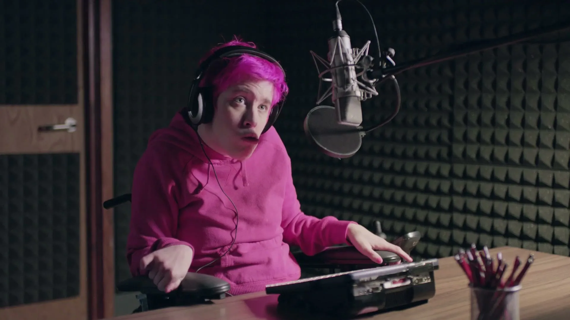 A person with bright pink hair and a pink hoodie sits in a wheelchair at a desk with a microphone in a sound booth.
