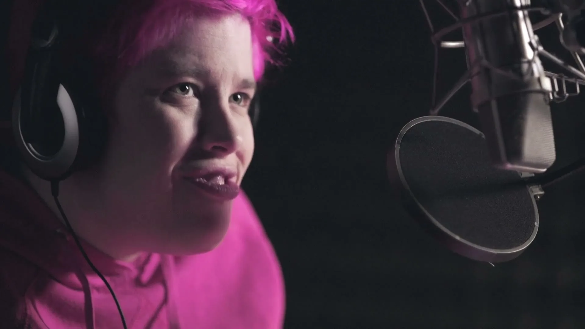 A young person with bright pink hair and headphones speaks into a microphone in a recording studio.