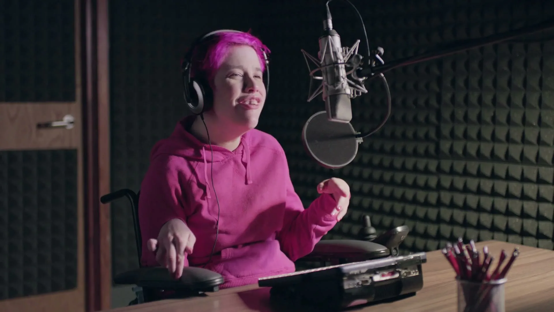 A young woman with pink hair and a disability, wearing headphones, sits in a wheelchair at a desk with a microphone in a recording studio.