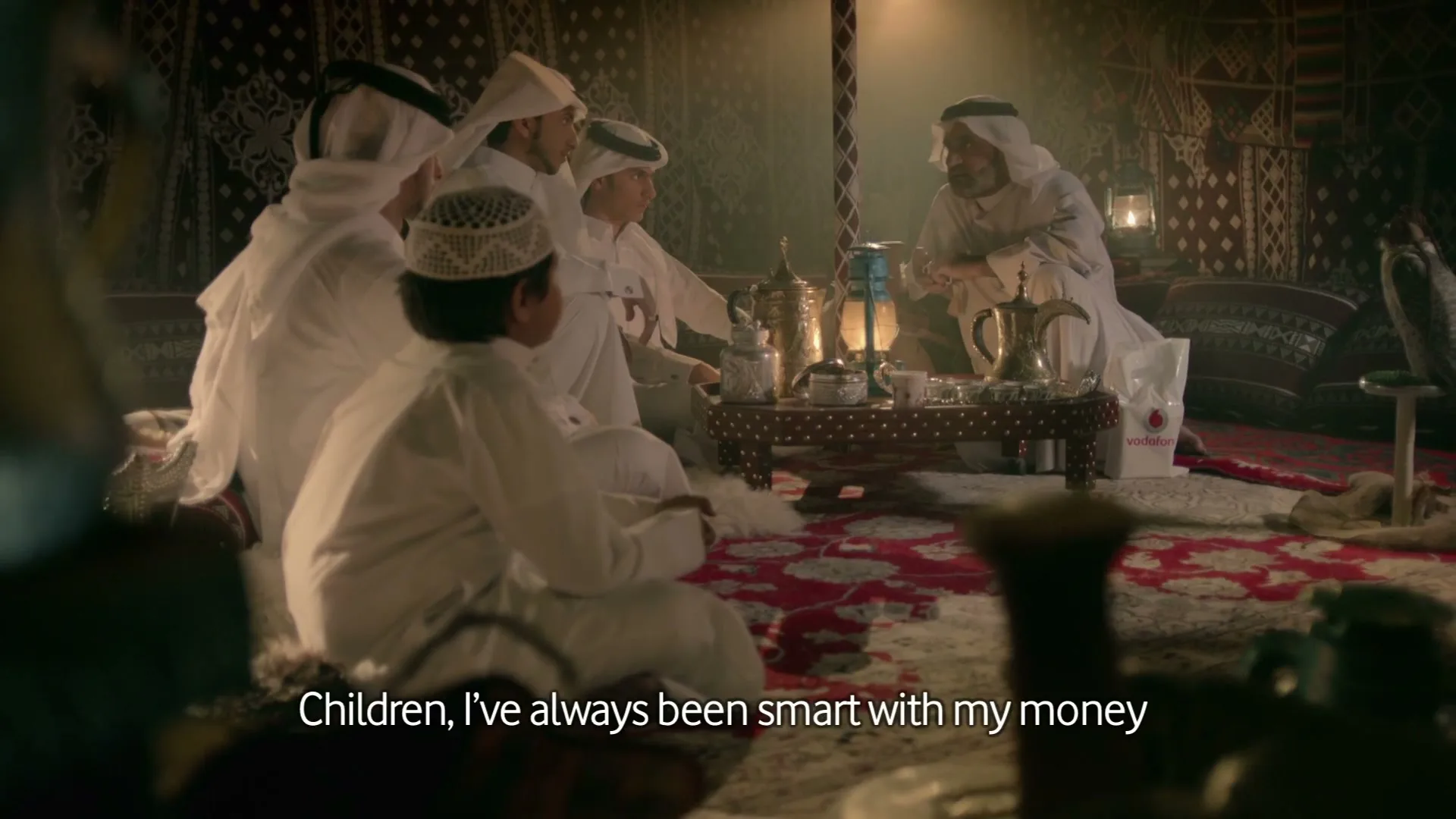 An older man in traditional white robes sits on a rug, talking to four younger men and boys, also in white robes, in a dimly lit tent.