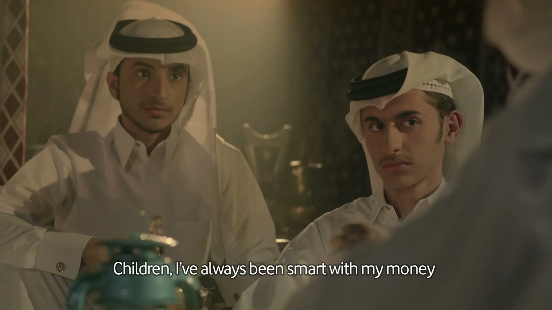 Two young men in traditional white thobes and ghutras listen intently to an unseen speaker in a dimly lit, traditional Arabic interior.