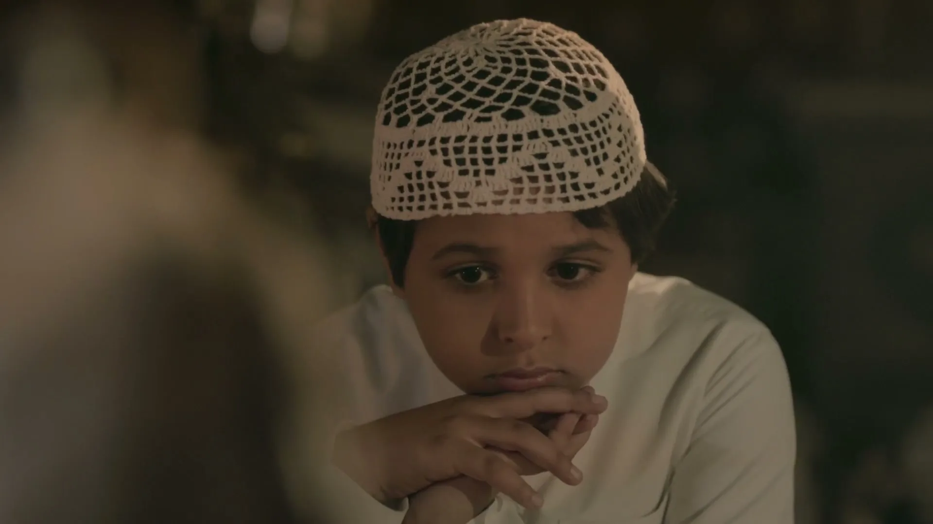 A young boy in a white kufi cap looks down thoughtfully, listening to someone off-screen in a dimly lit interior.