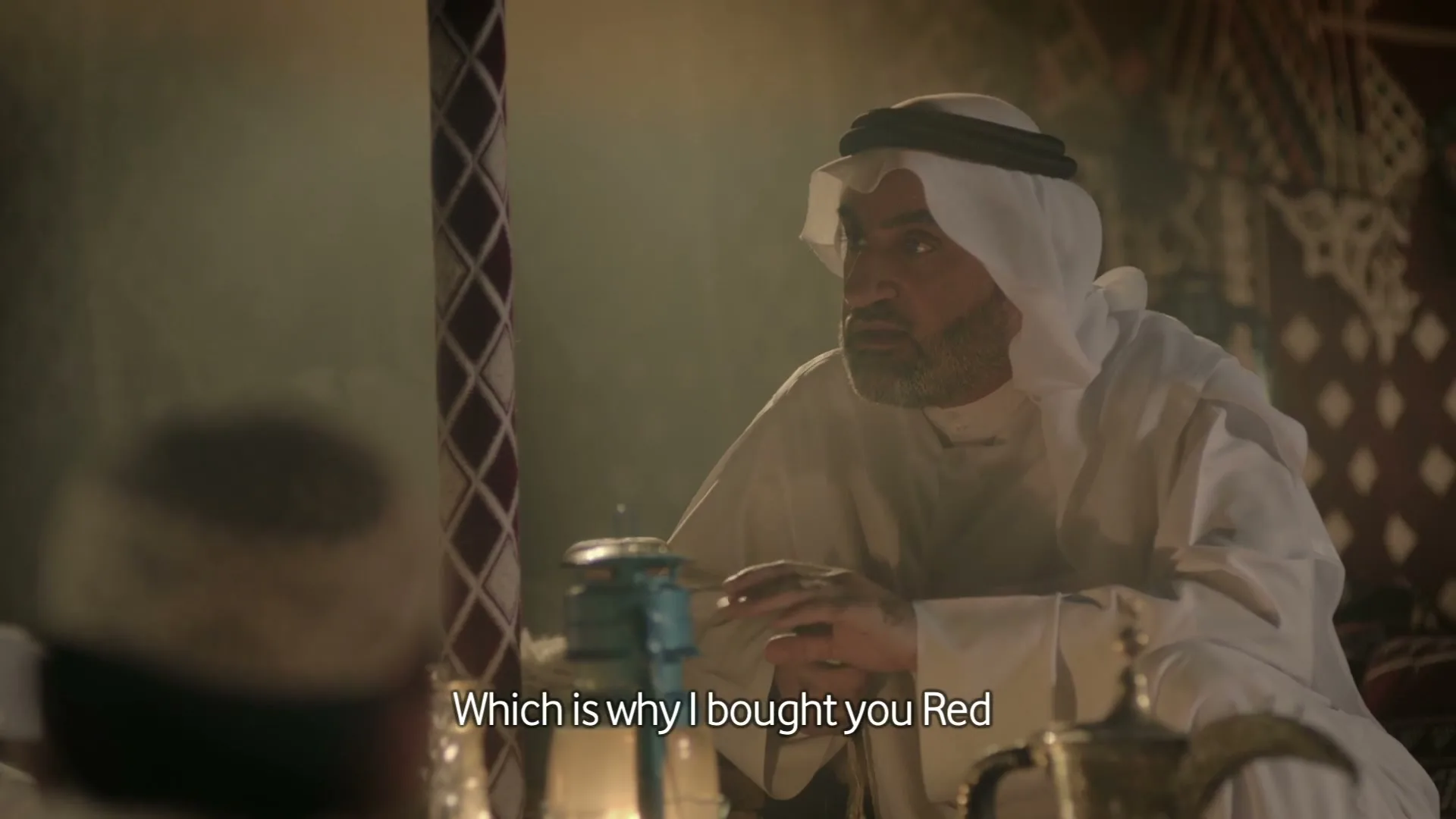 An older man in traditional Arab attire sits in a tent, gesturing with his hands while speaking to someone off-screen.