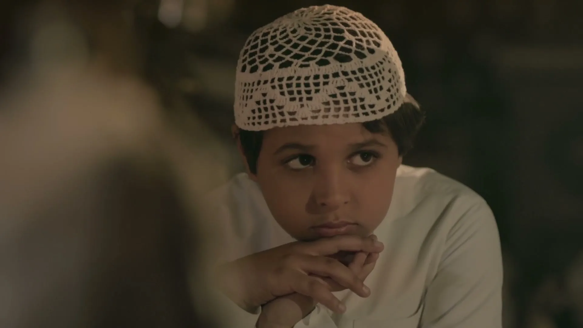 A young boy in a white crocheted cap and white shirt looks up with a serious expression, listening intently to someone off-screen in an interior setting.