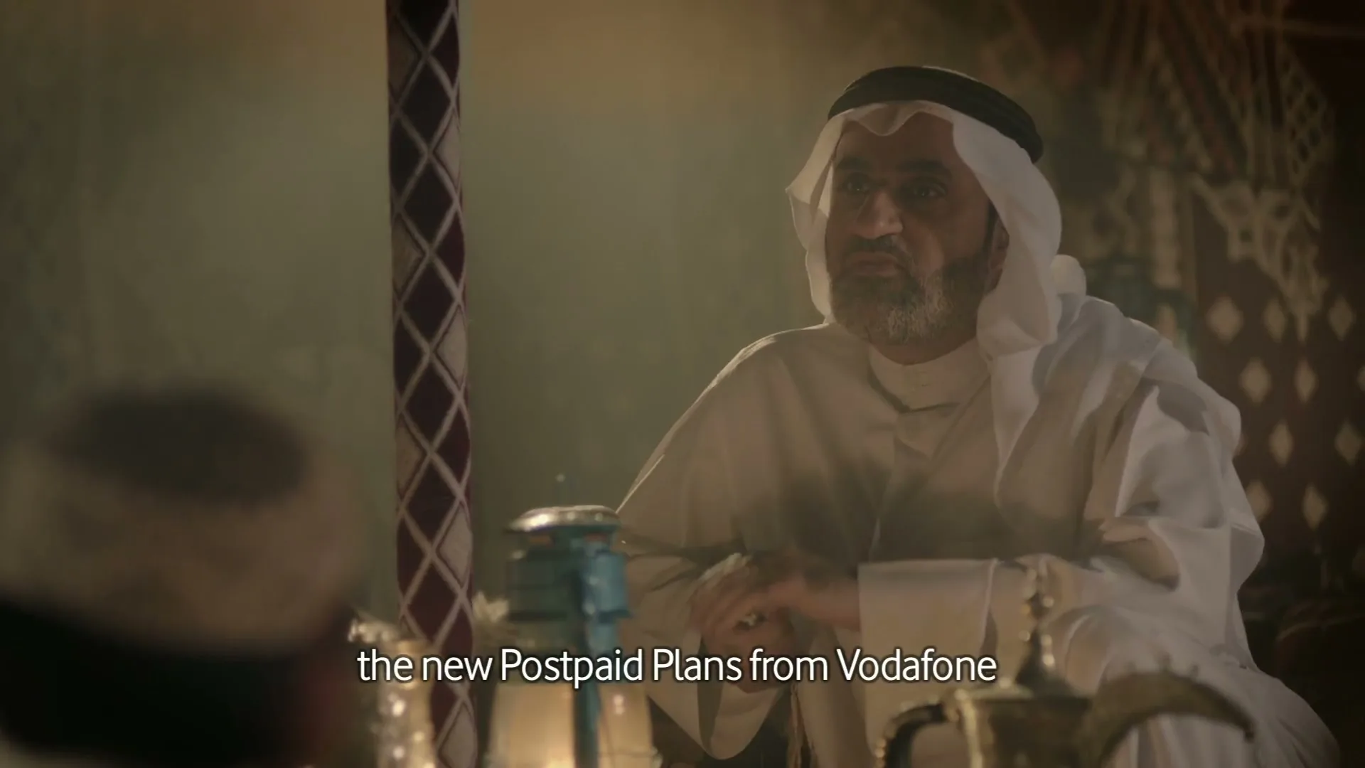 A man in traditional Arab attire sits in a tent, gesturing while speaking to someone off-camera, with warm lighting illuminating the scene.