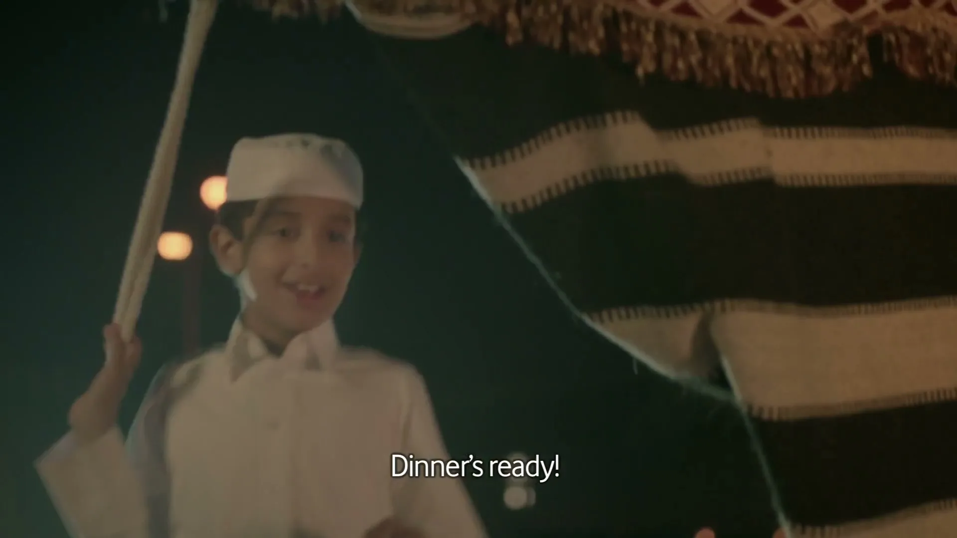 A young boy in traditional white attire smiles and waves, calling out "Dinner's ready!" from behind a striped tent at night.