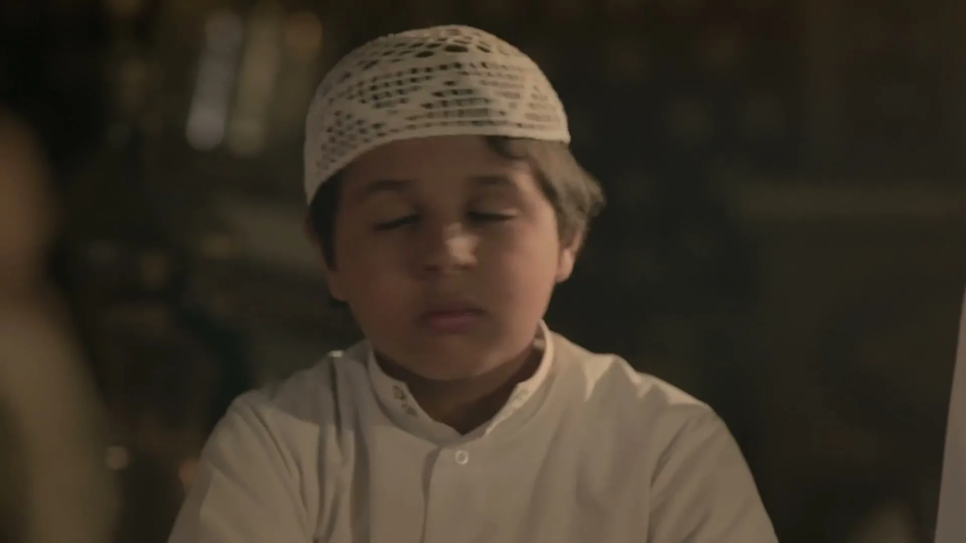 A young boy in a white prayer cap and shirt looks around, then closes his eyes, then looks to the side, at a dinner table.
