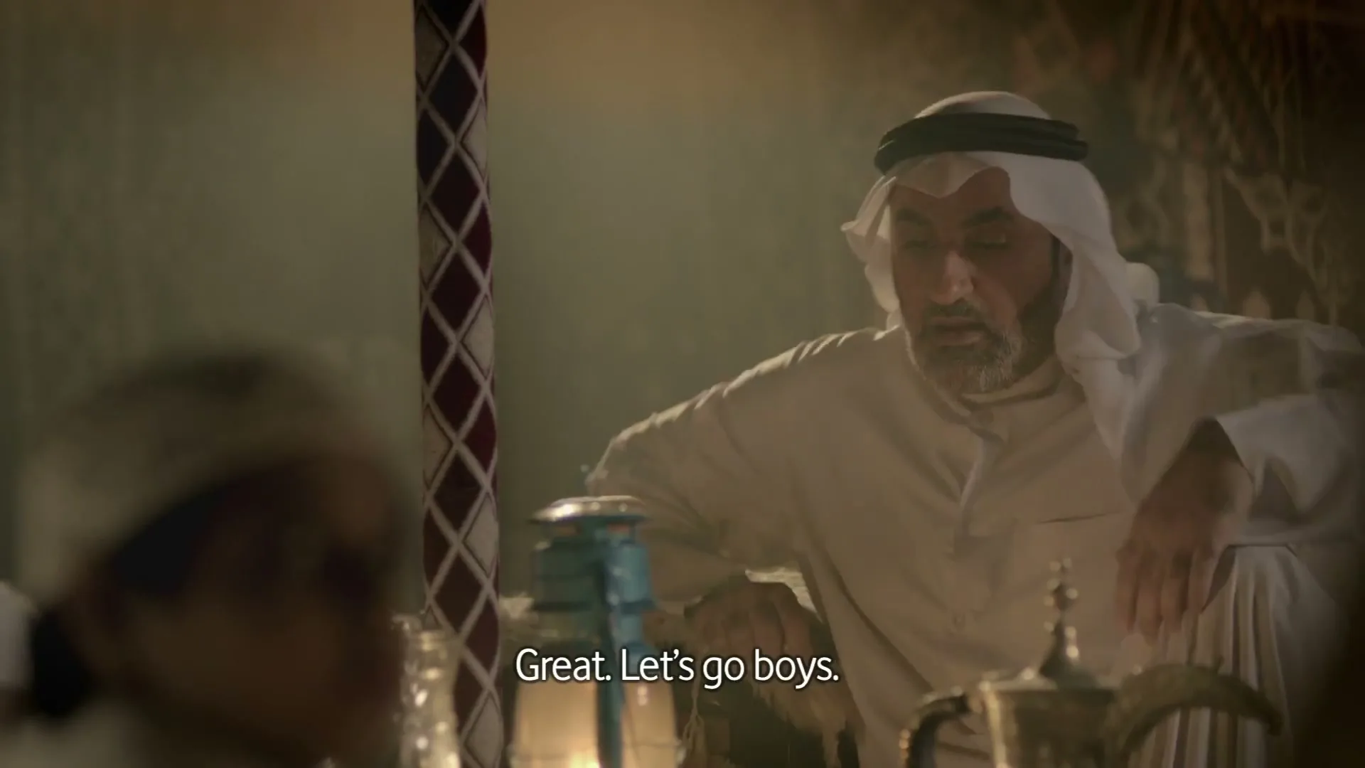 A man in traditional Arab attire speaks to someone off-camera, while sitting in a tent with a lantern and a coffee pot.
