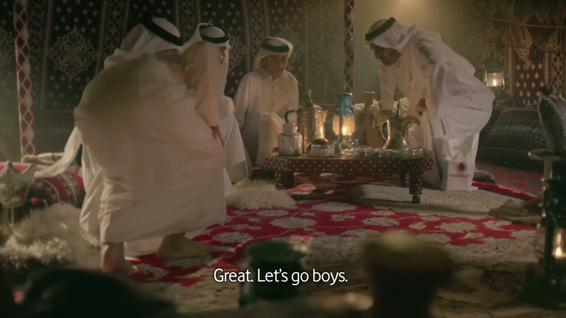 A group of men in traditional white thobes and ghutras gather around a low table with drinks and a lantern inside a tent, as one man stands up.