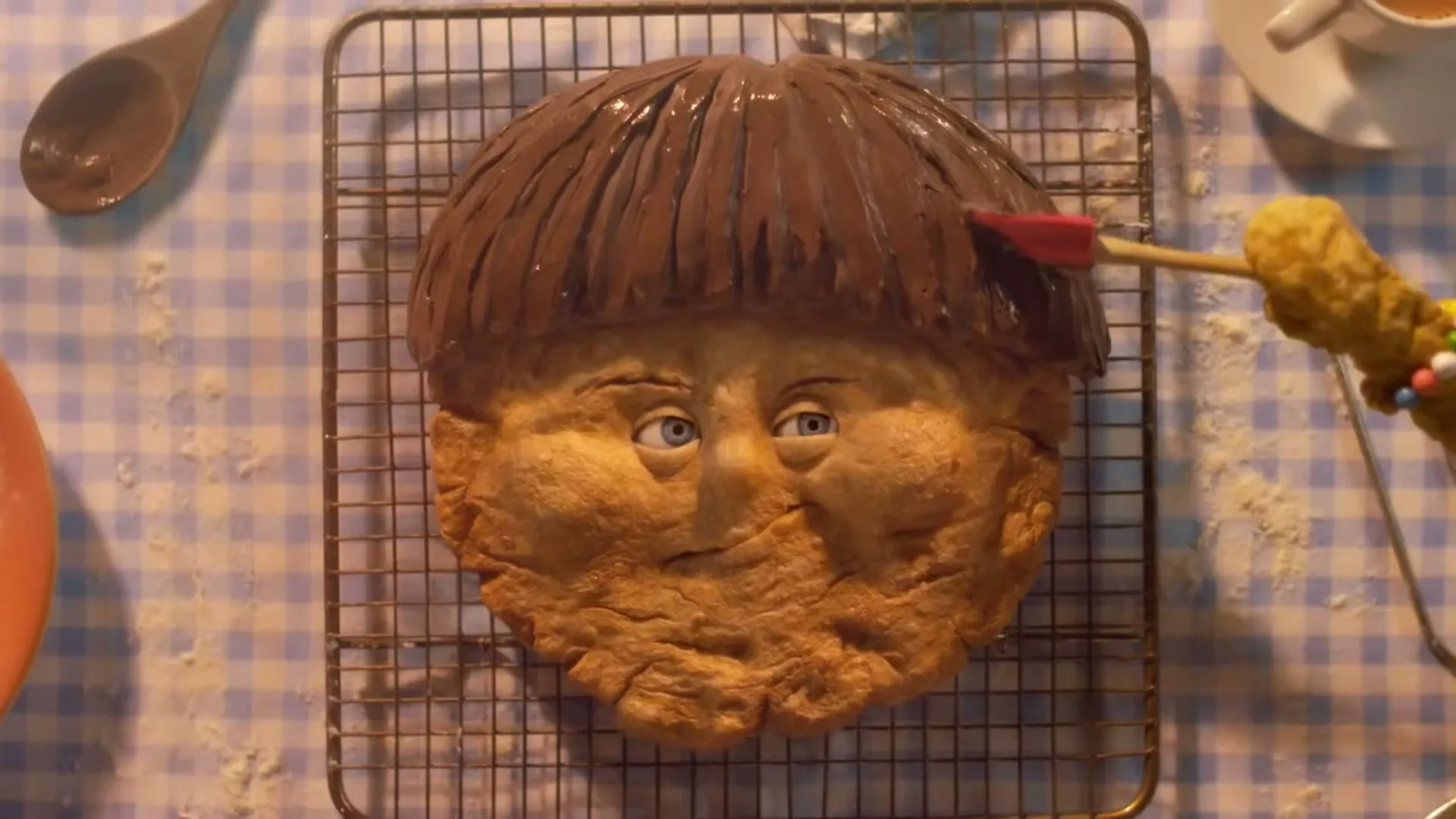 A stop-motion animated biscuit boy with chocolate hair is being brushed by a yellow-gloved hand on a cooling rack on a blue and white checkered tablecloth.