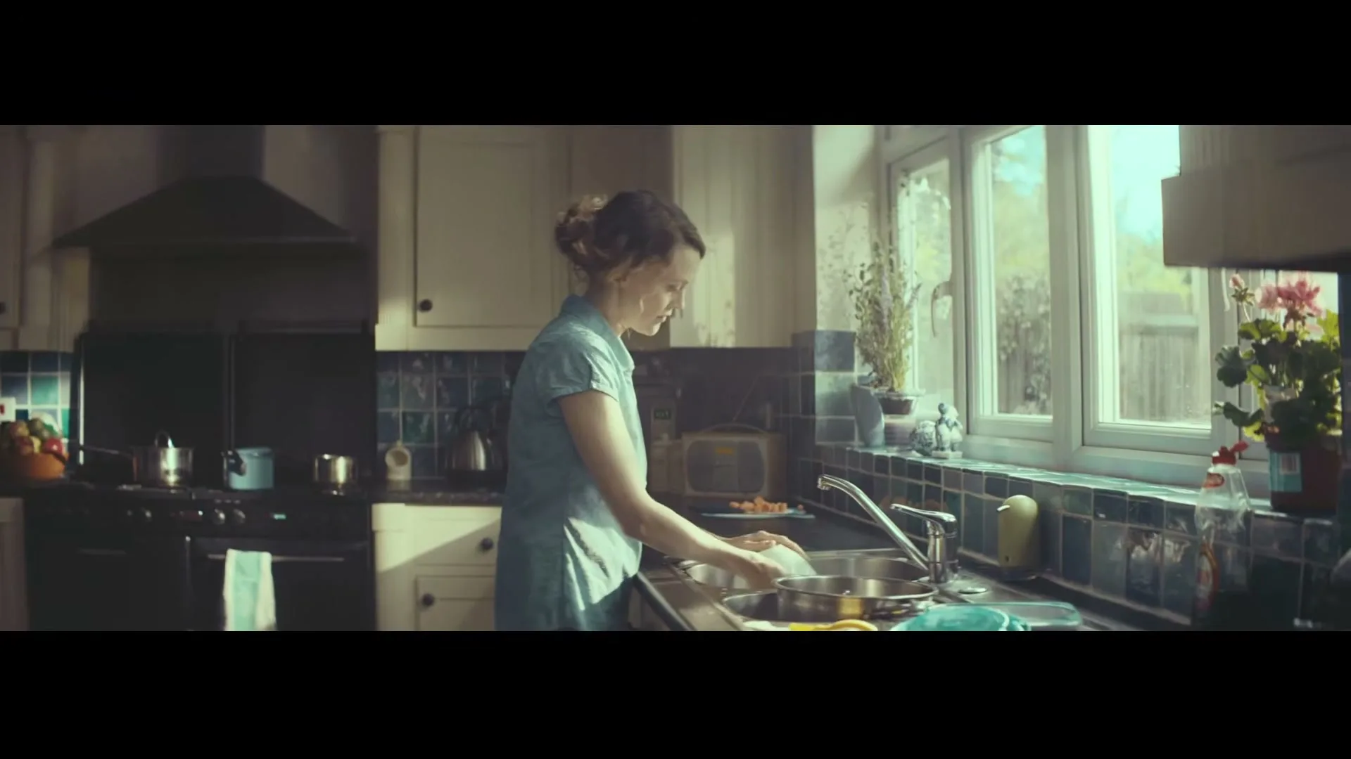A woman with her hair tied back, wearing a light blue dress, washes dishes at a sink in a sunlit kitchen.