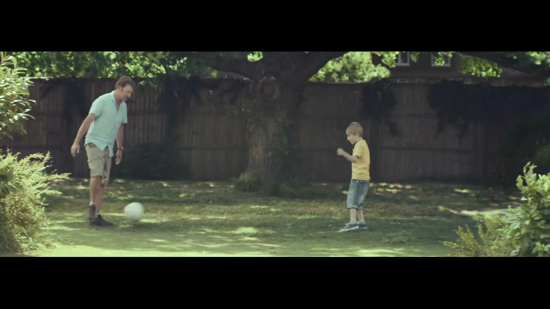 A father and son play soccer in their backyard on a sunny day, with the father wearing a light blue shirt and shorts, and the son in a yellow shirt and shorts.