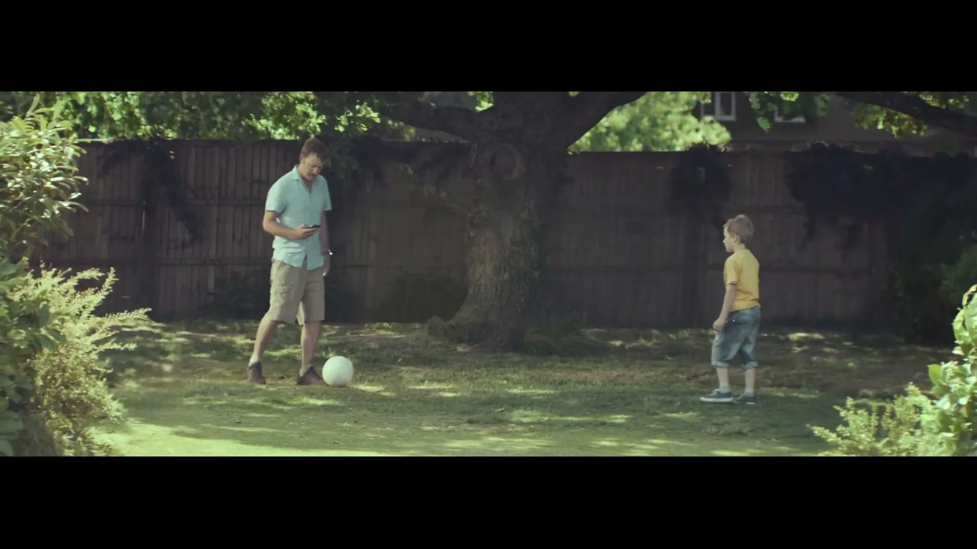 A father and son play soccer in a sunny backyard, but the father gets distracted by his phone, leaving the boy to kick the ball alone.