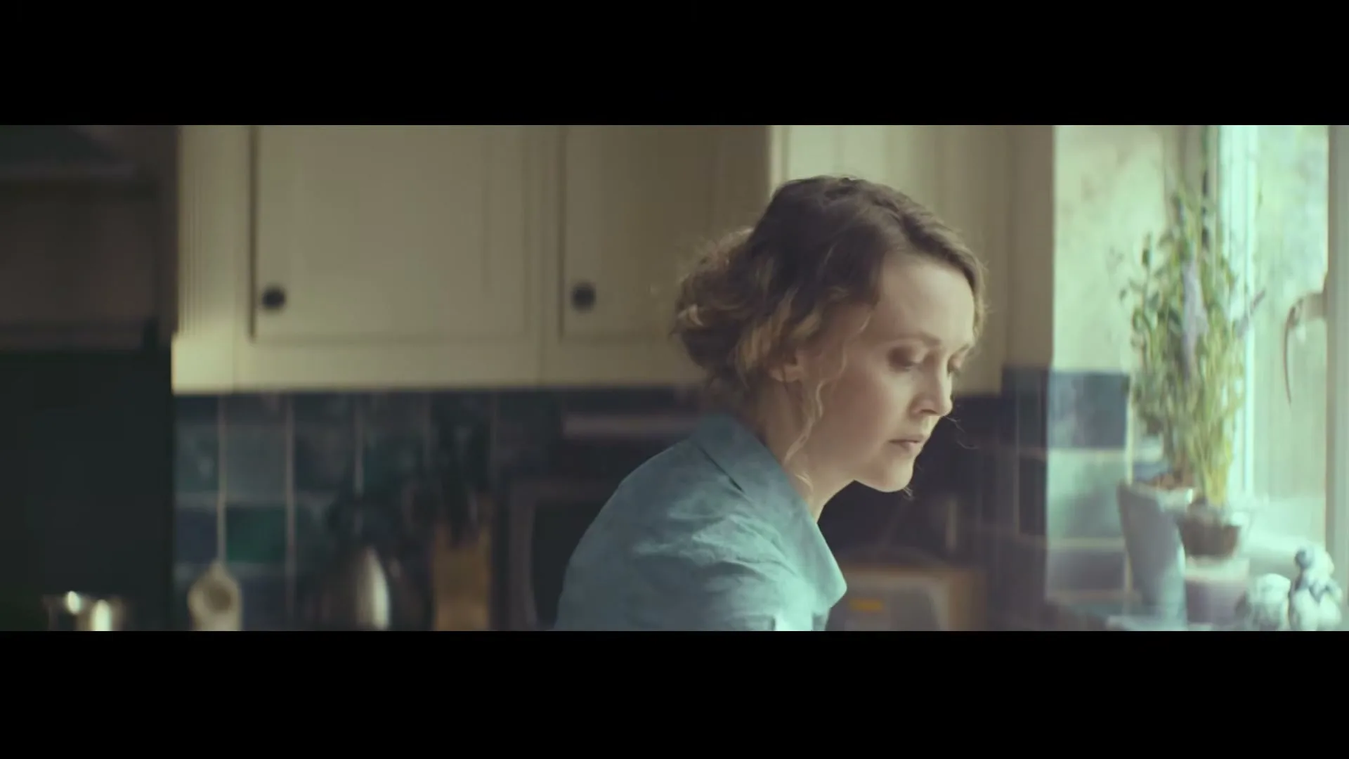 A woman with curly hair, wearing a light blue shirt, is seen in profile, looking down while doing dishes in a kitchen.