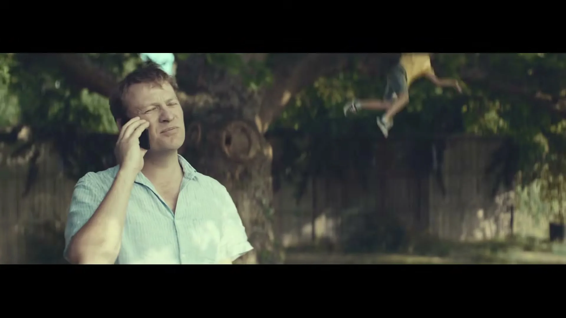 A man in his 30s talks on the phone, looking concerned, while a boy swings in the background of a sunny backyard.