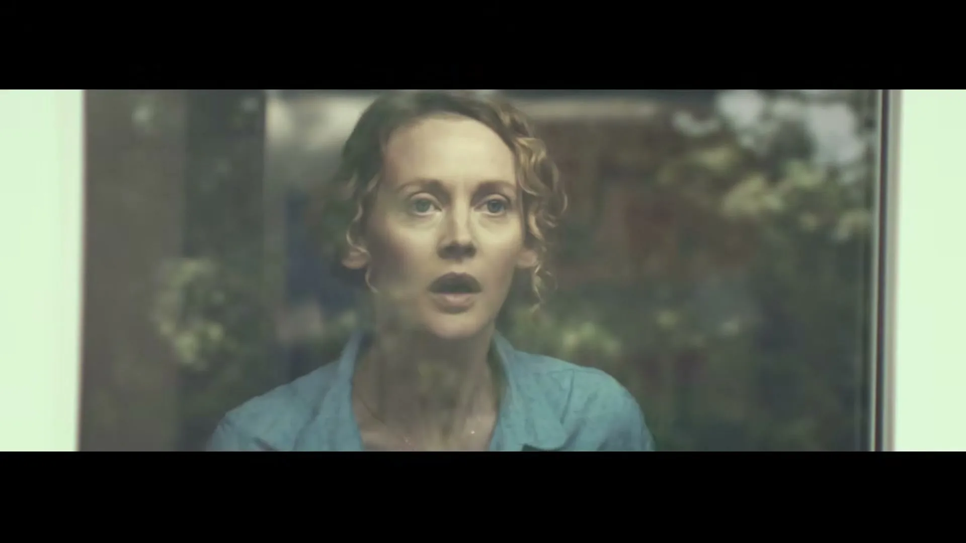 A woman with curly blonde hair and a blue shirt looks out a window with a worried expression.