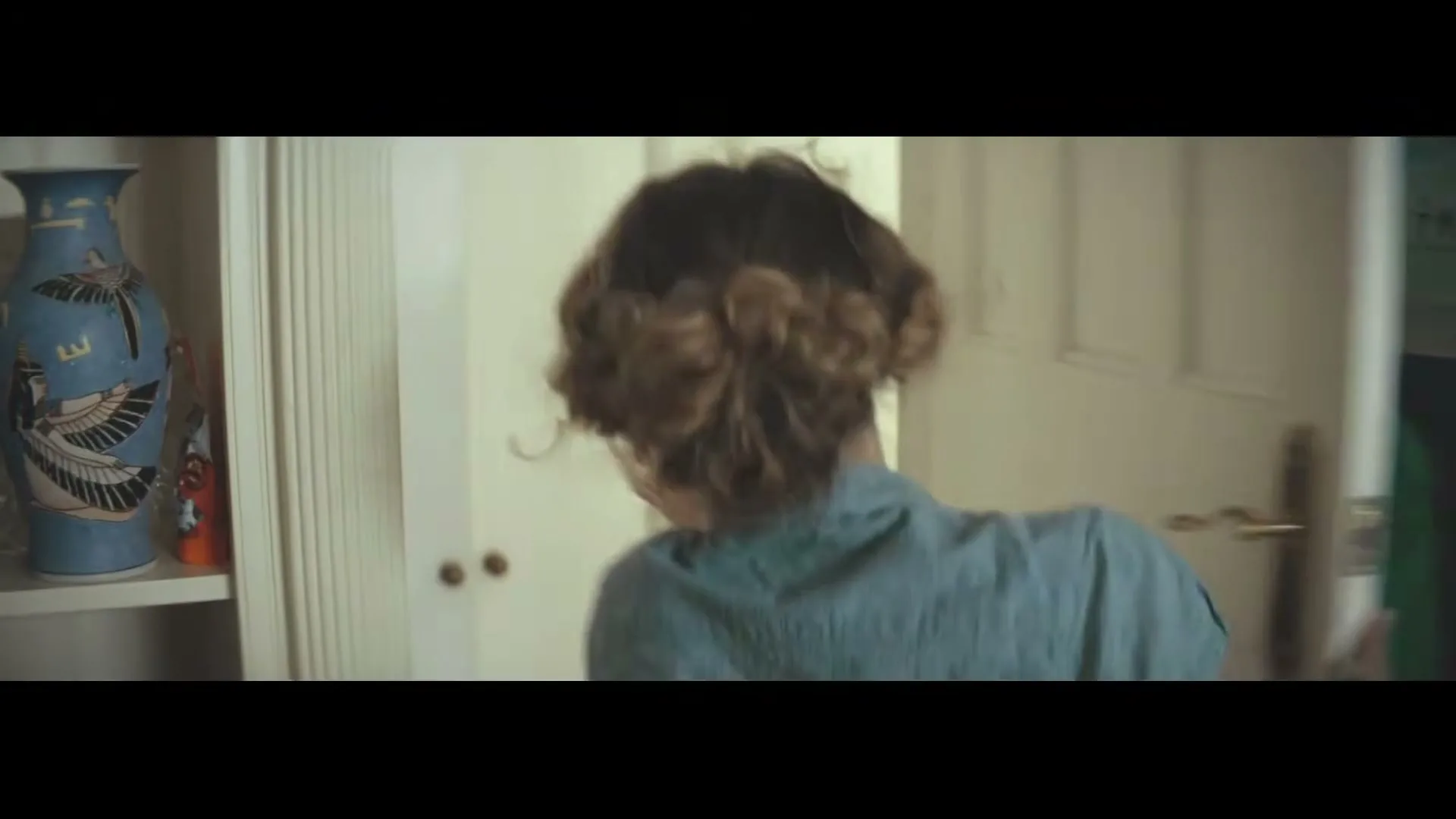 A woman with curly hair rushes through a home, grabbing a green and black St John Ambulance jacket from a door, preparing to help.