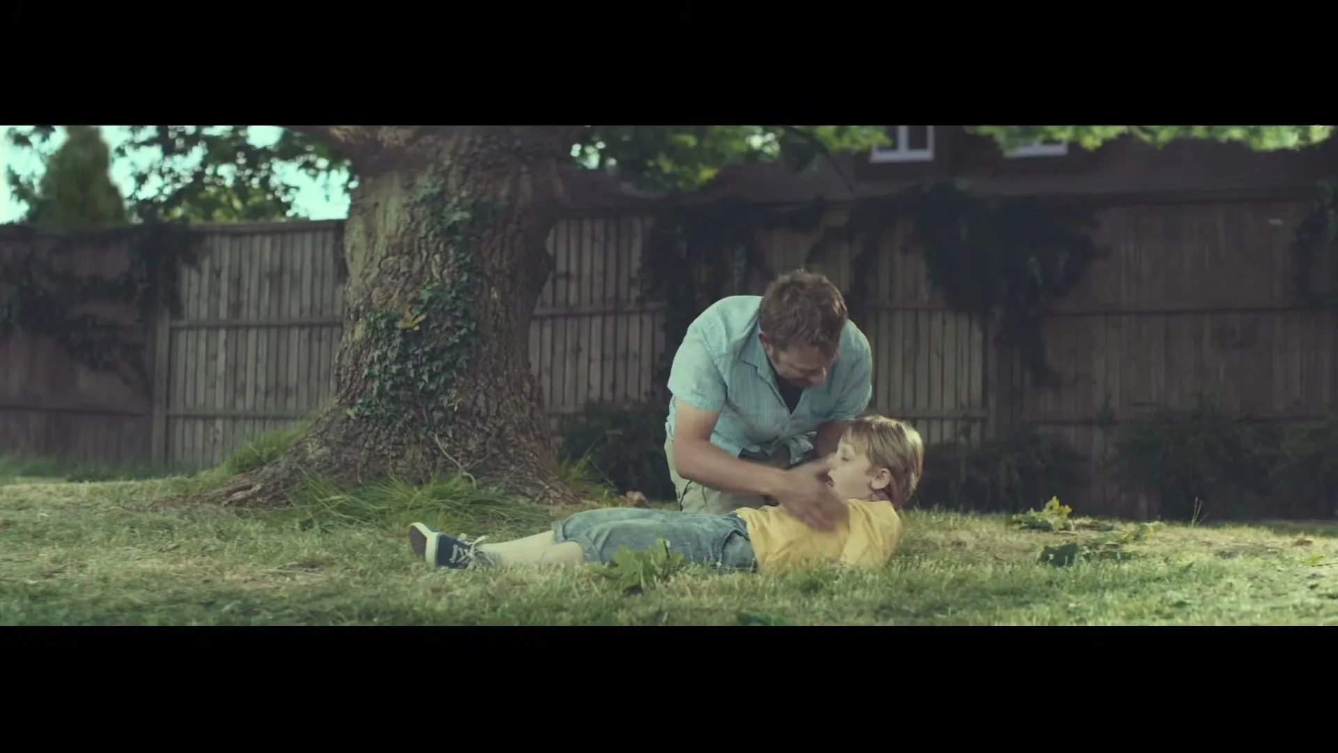 A man in a light blue shirt kneels over a young boy in a yellow shirt and blue shorts, who is lying on the grass in a backyard.