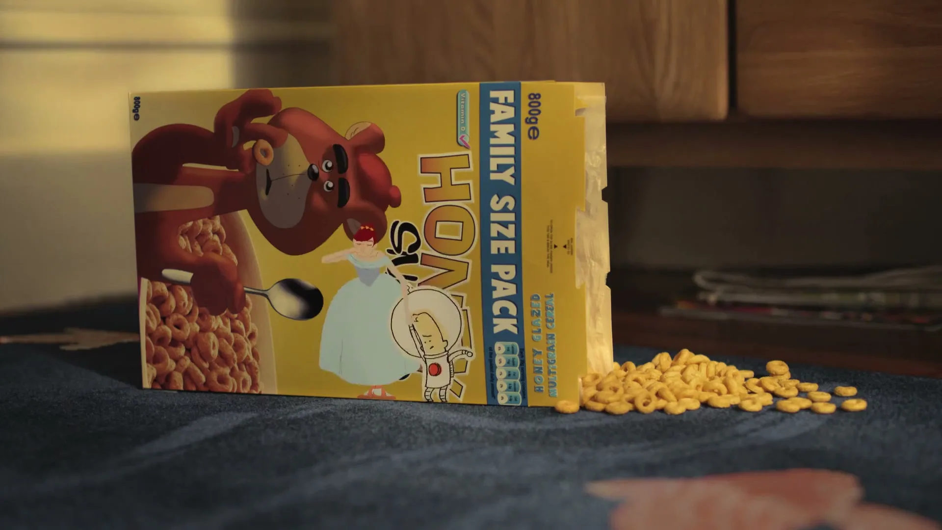A box of "Honey Snacks" cereal with a cartoon bear and princess on the front, sits on a blue rug with spilled cereal, in a living room.