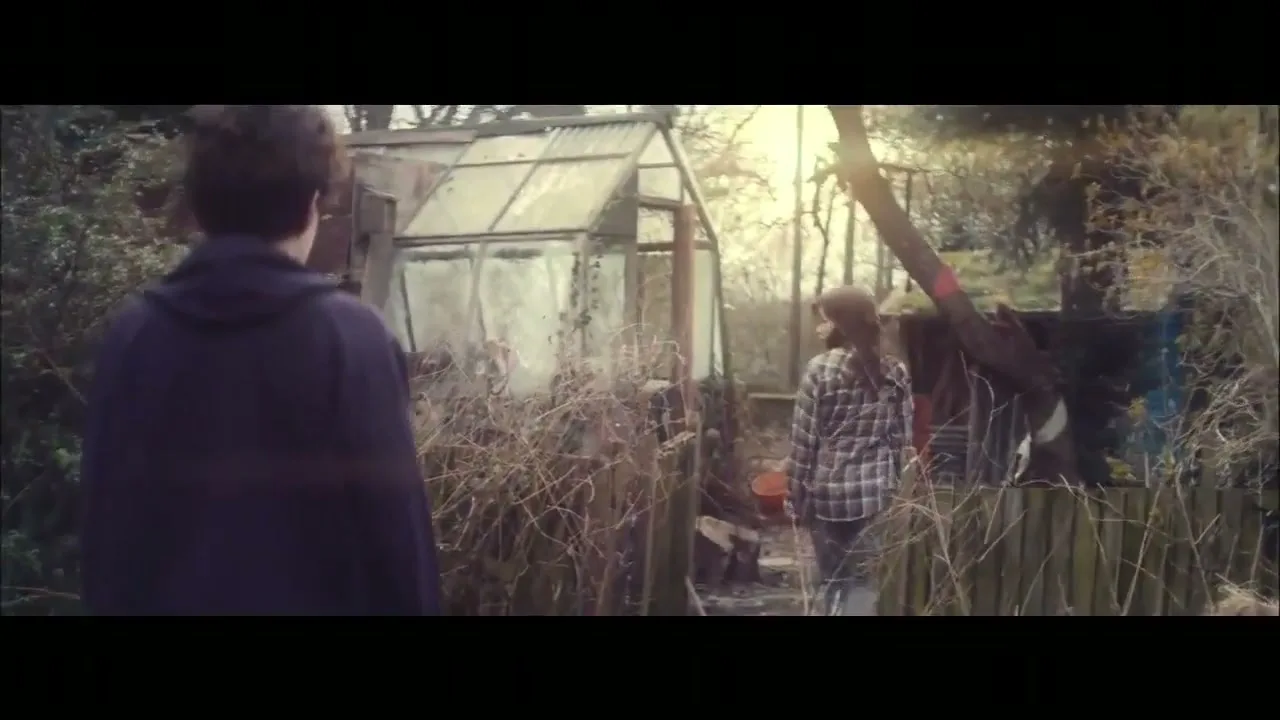 A young man in a dark hoodie watches a young woman in a plaid shirt walk away from him towards a greenhouse in a overgrown garden.