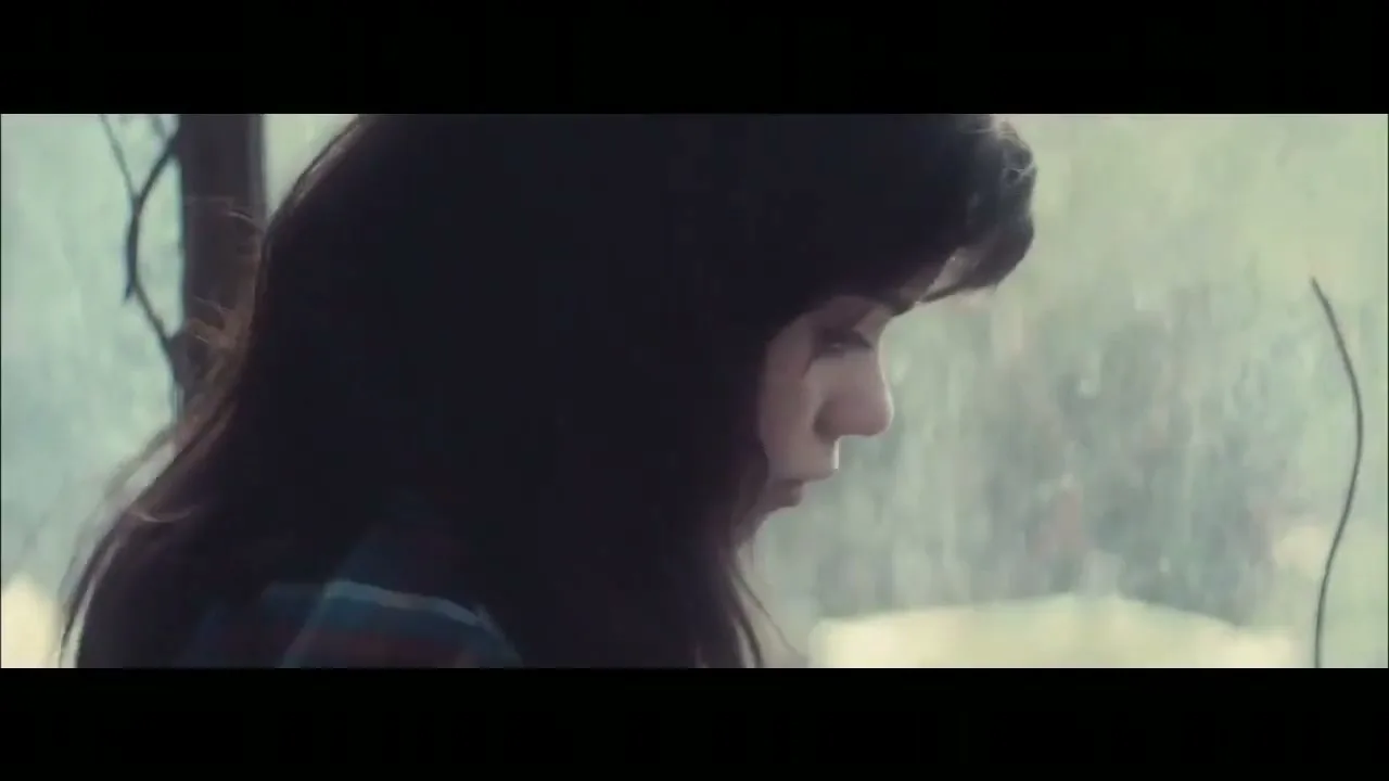 A young woman with dark hair, wearing a plaid shirt, looks down with a pensive expression in a dimly lit room with a window in the background.