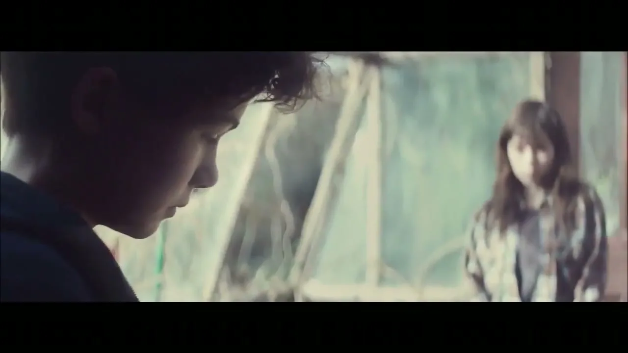 A young boy with curly hair looks down, while a girl with long hair stands blurred in the background, in a greenhouse.