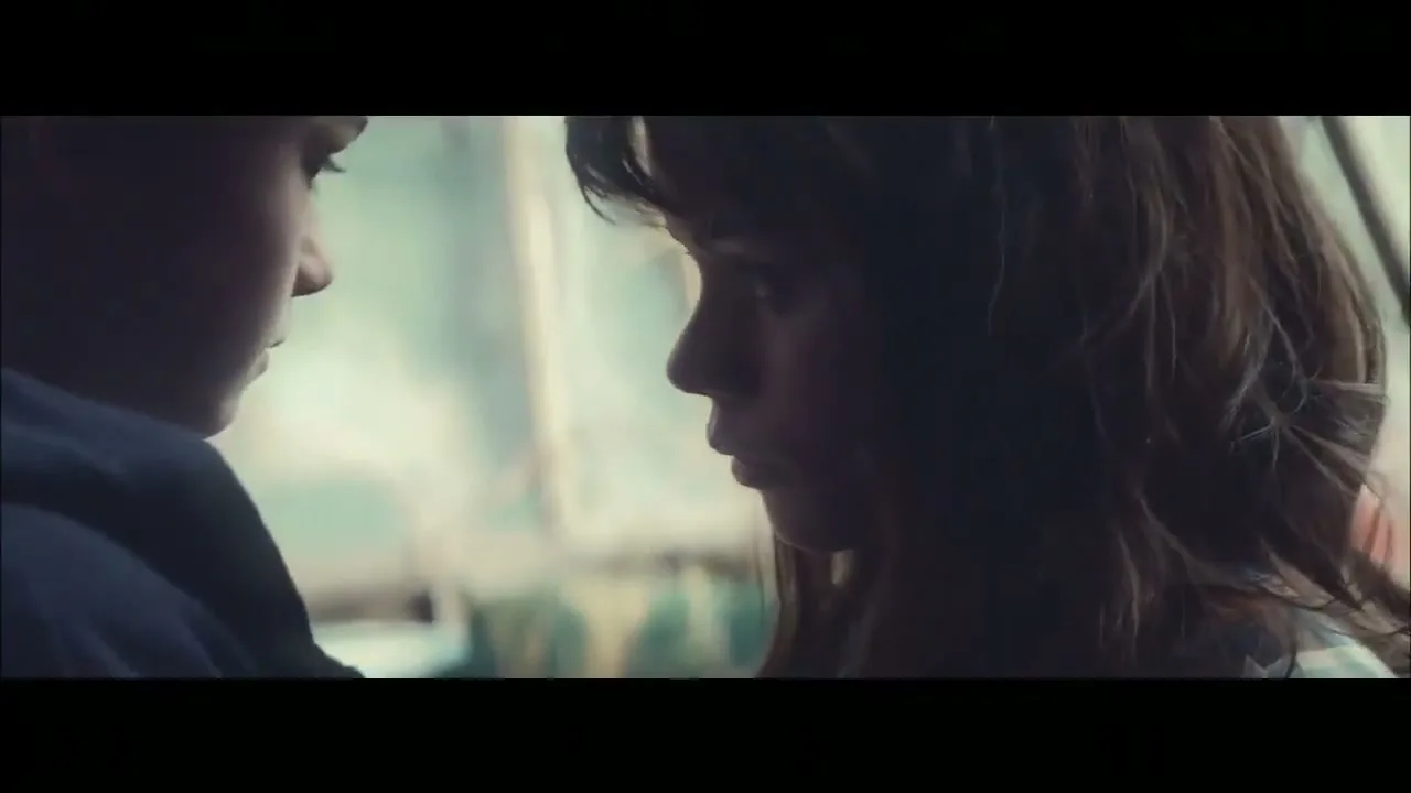 A young boy and girl stand close, looking at each other with serious expressions, in a softly lit, blurry outdoor setting.