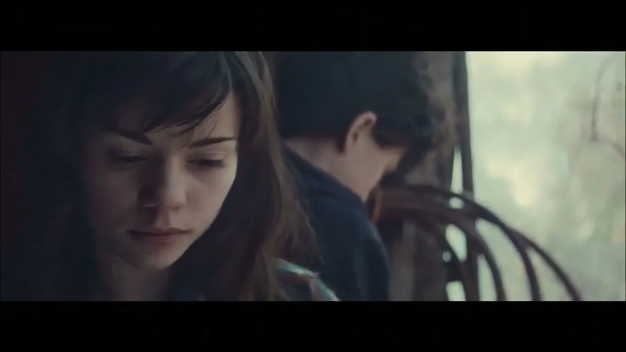 A young woman with dark hair looks down thoughtfully, while a young man in a dark shirt is visible behind her, looking out a window on a train.