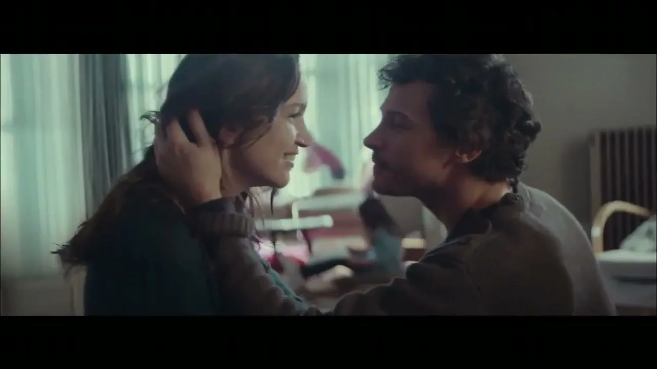 A young couple, a woman with long dark hair and a man with curly dark hair, kiss passionately in a softly lit room.