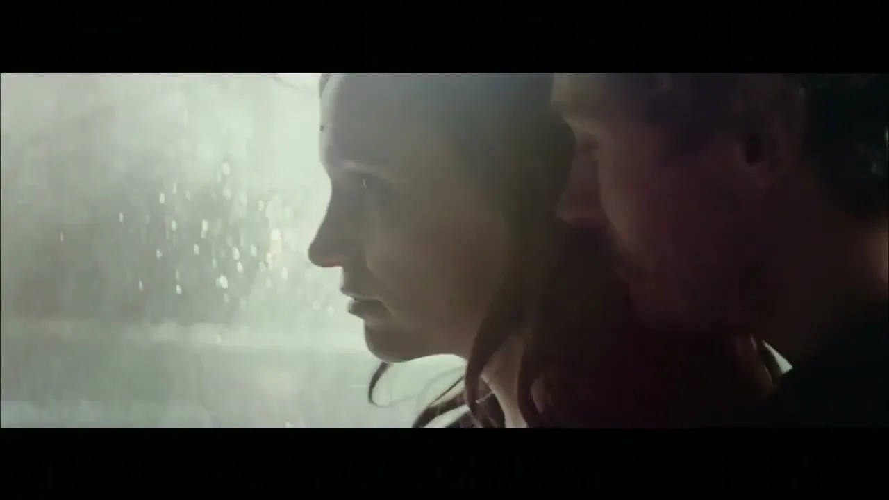 A young couple, a woman with long brown hair and a man with a beard, stand close together, looking at each other in a dimly lit room.