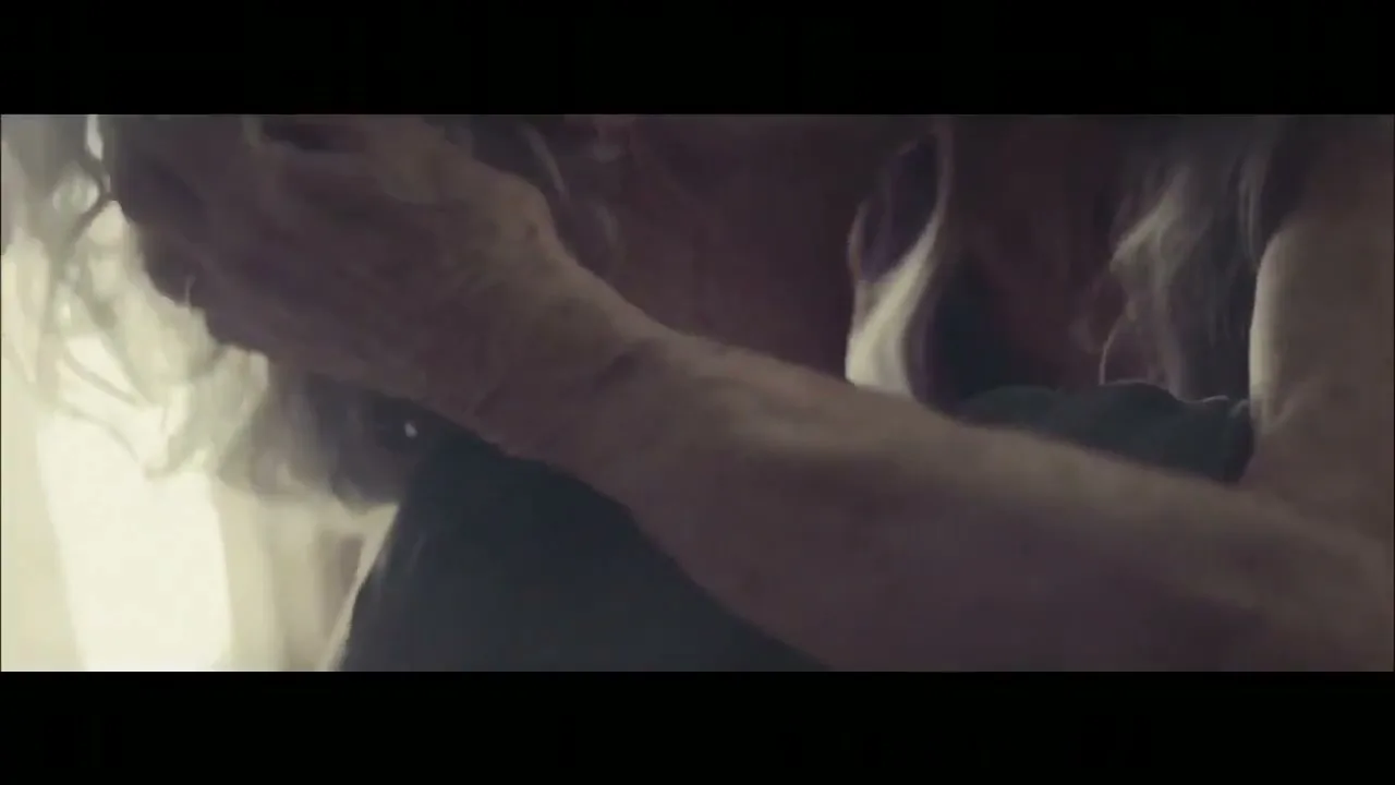 An elderly couple, a man and a woman, embrace intimately, their faces close together, in a soft, naturally lit interior setting.