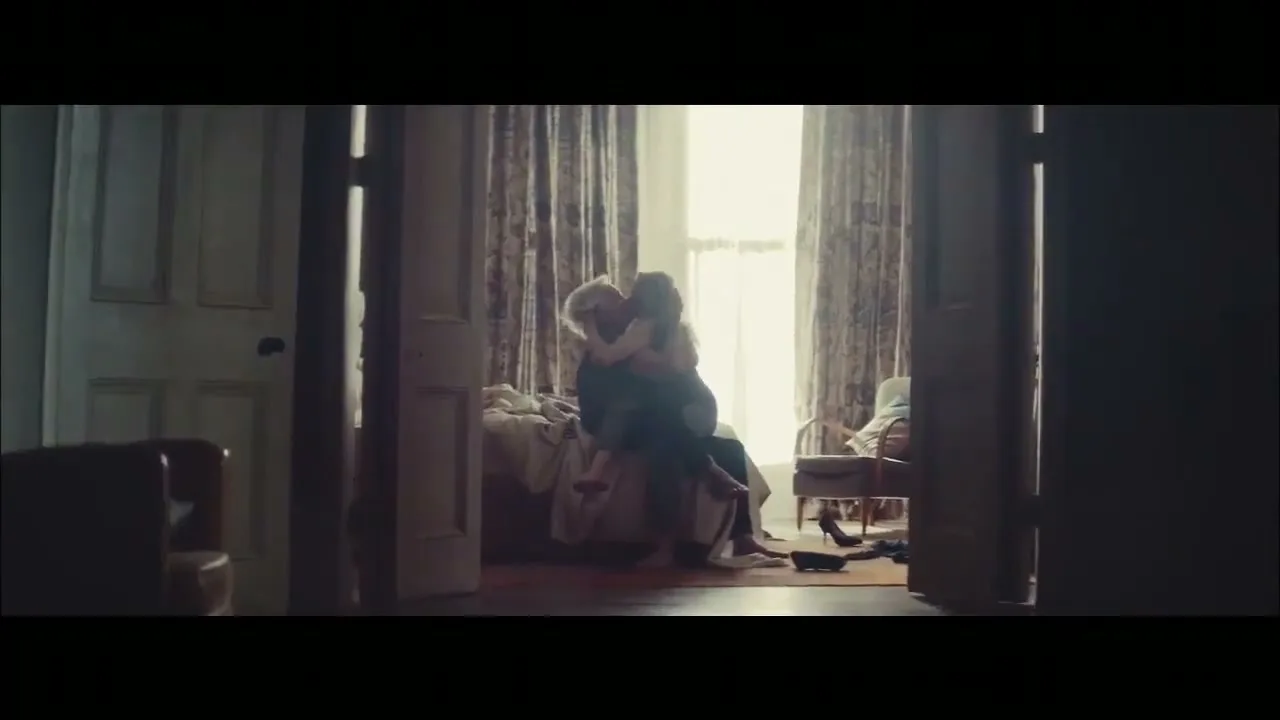 A man and woman embrace passionately on a bed in a sunlit bedroom, viewed through a doorway from a dark foreground room.