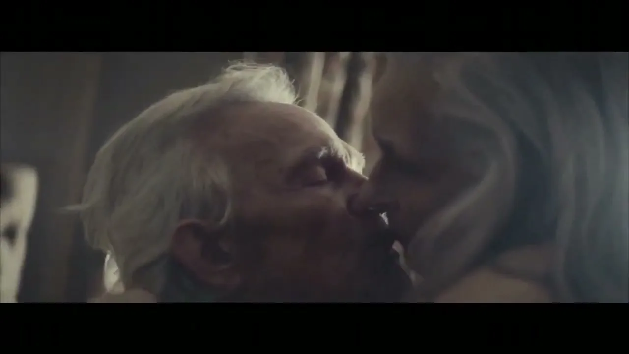 An elderly man with white hair and a woman with long blonde hair share a tender kiss, their faces close together in a dimly lit interior.