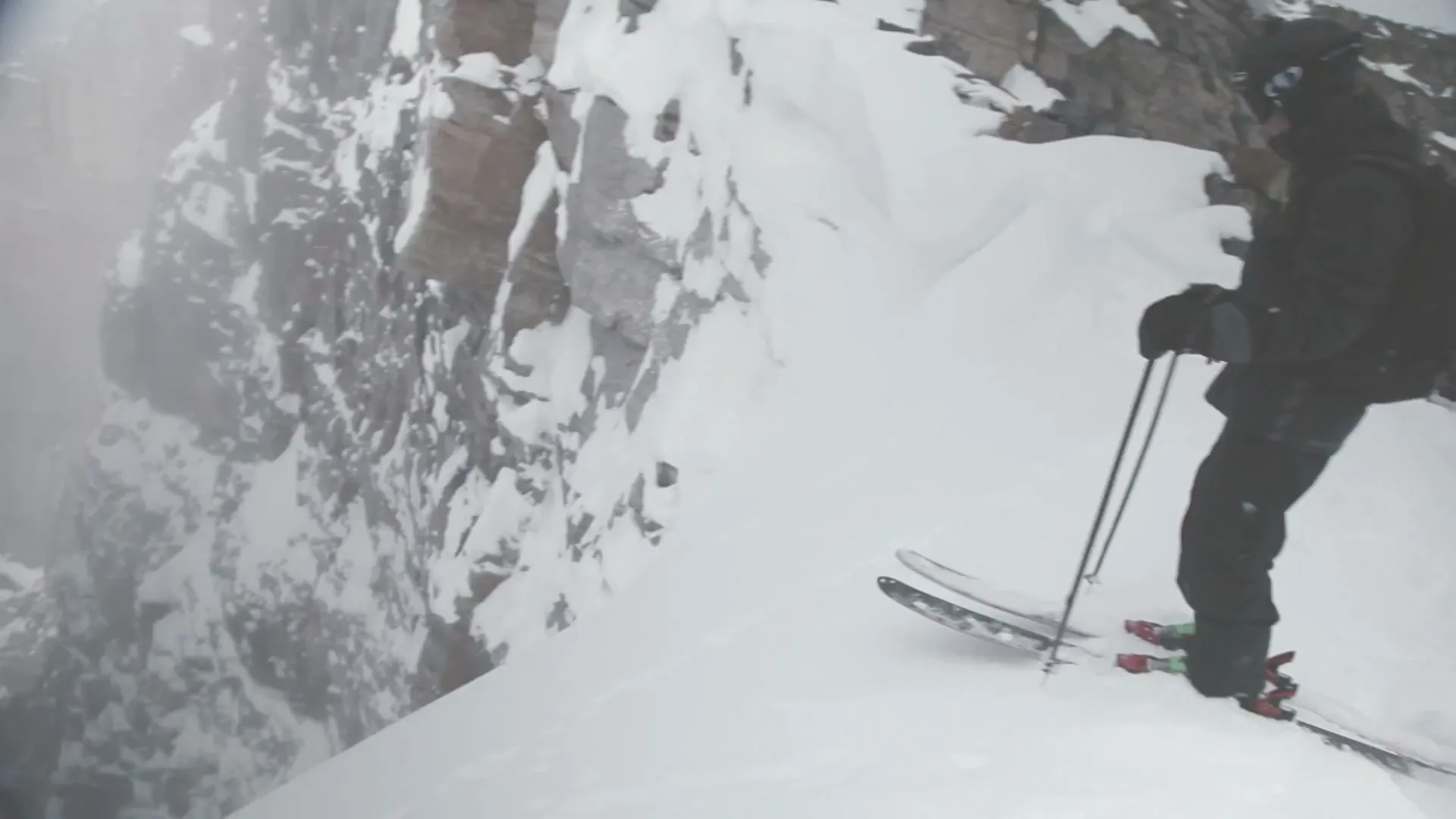 A skier in black winter gear stands on a snowy mountain ridge, looking down into a foggy abyss, preparing to descend.