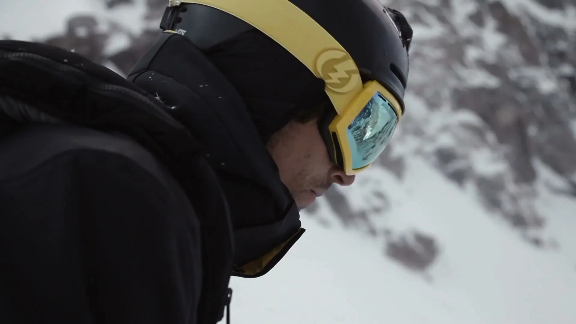 A male skier in a black jacket and yellow helmet looks down, preparing to ski on a snowy mountain.