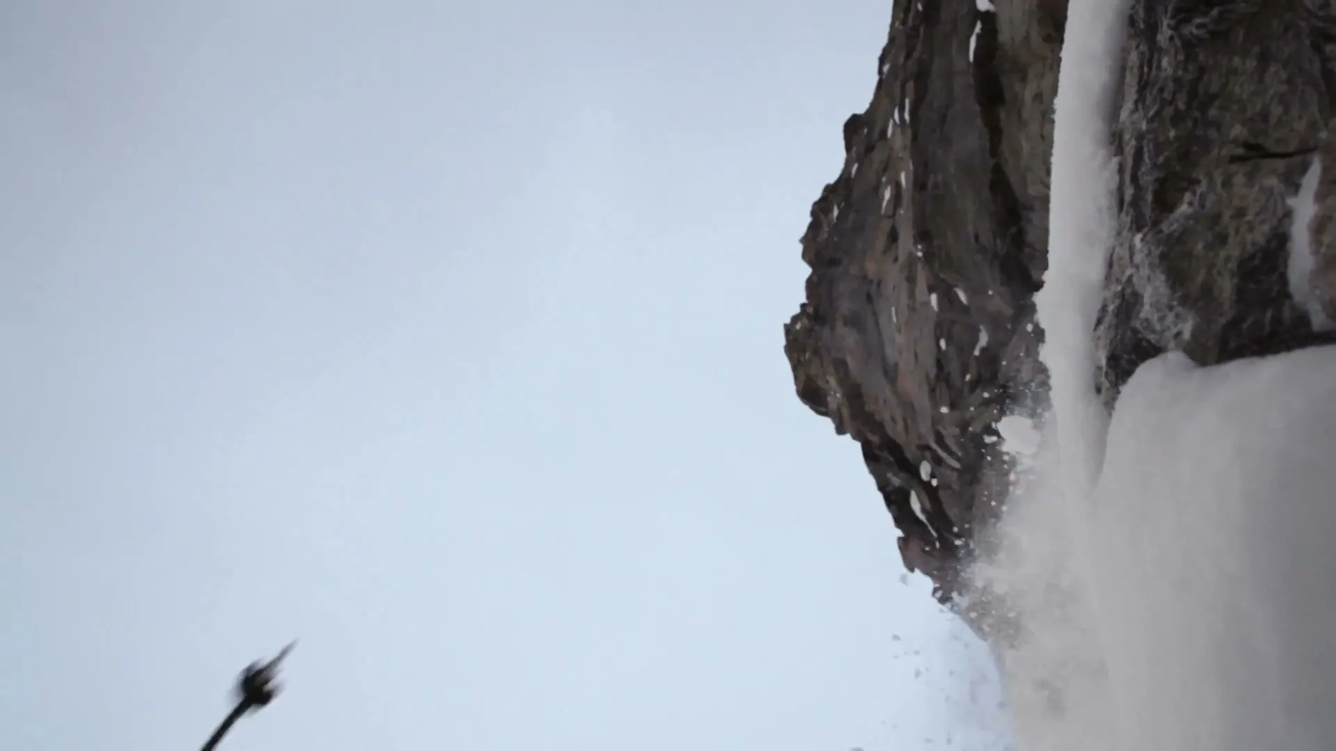 A male ice climber in winter gear is seen from a low angle, climbing a steep, icy rock face as snow and ice fall around him.