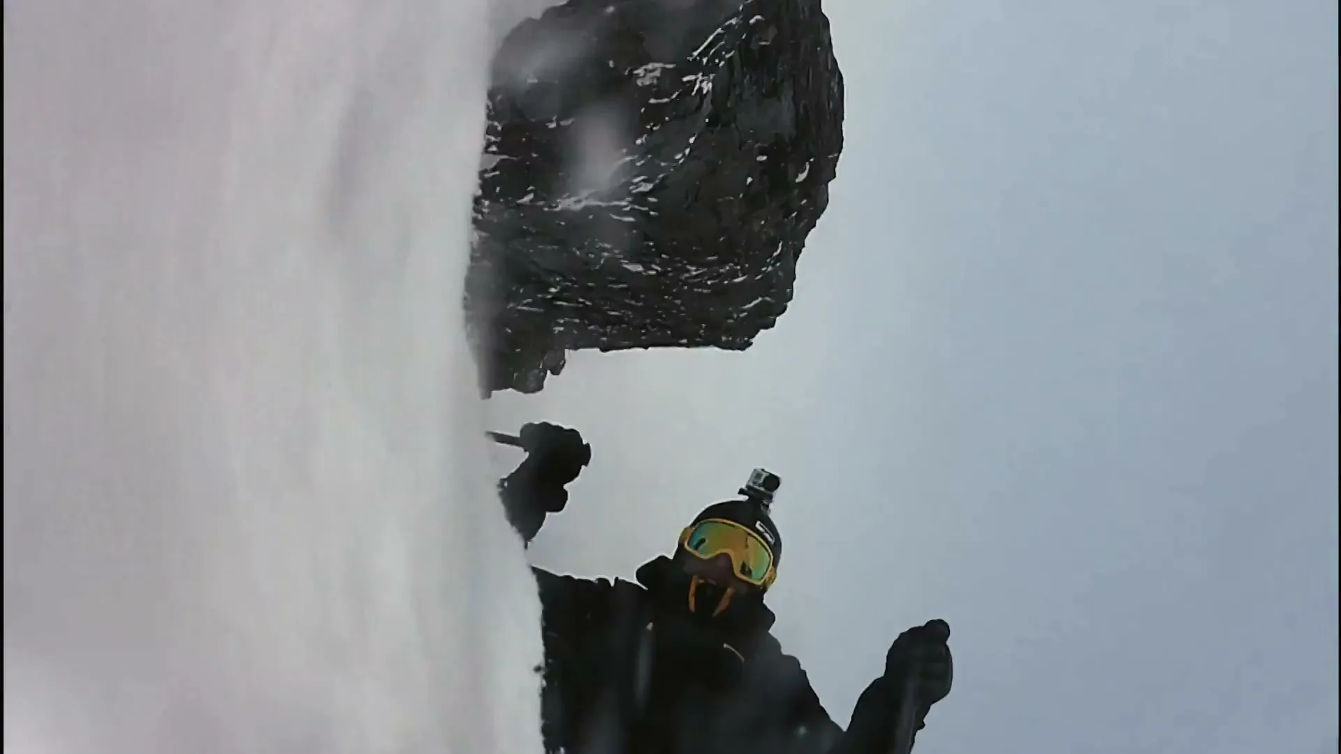 A skier in black gear and yellow goggles falls down a snowy mountain, captured from a POV angle.