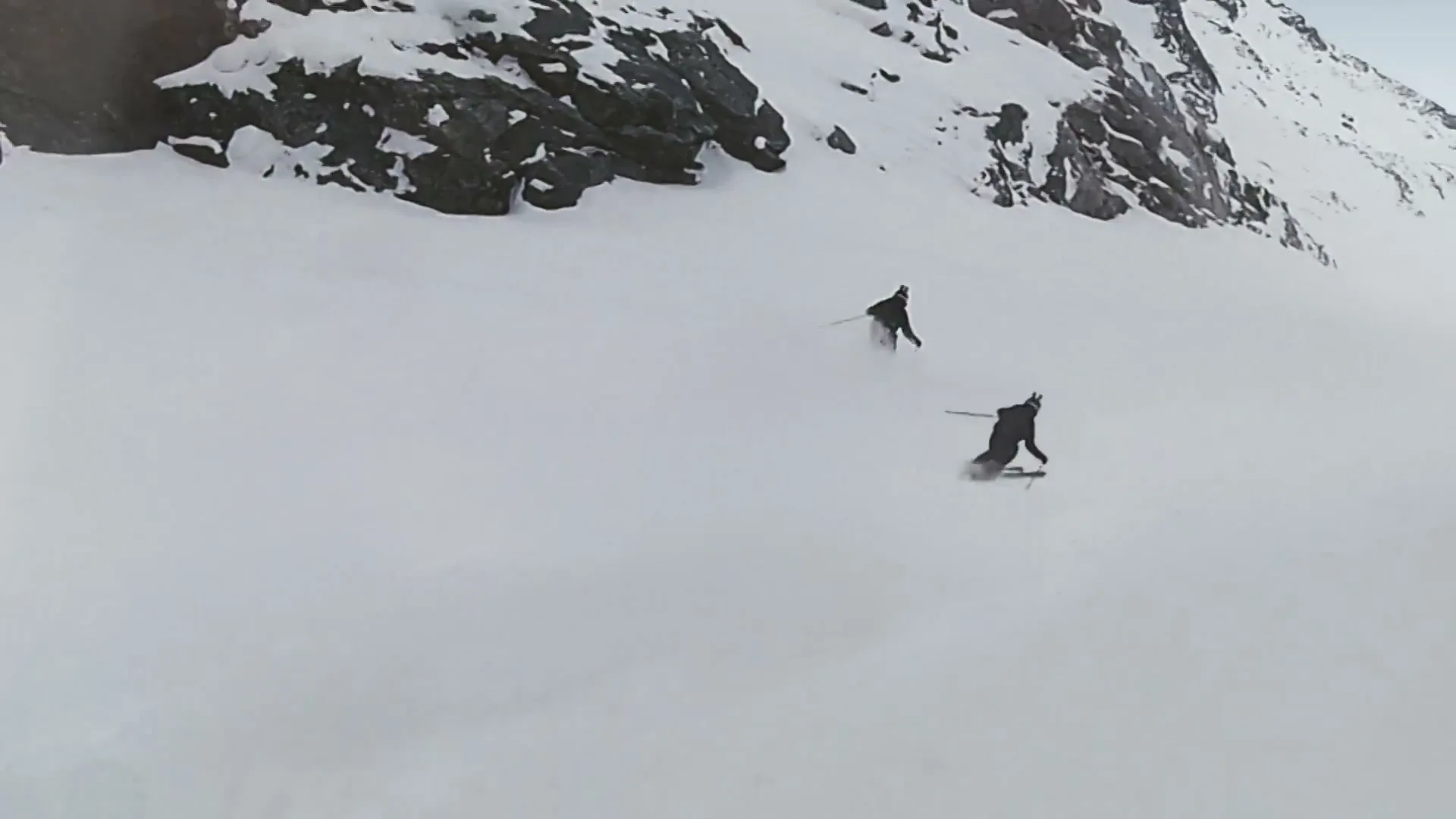 Two skiers in dark gear descend a vast, snow-covered mountain slope, kicking up powder as they carve turns.