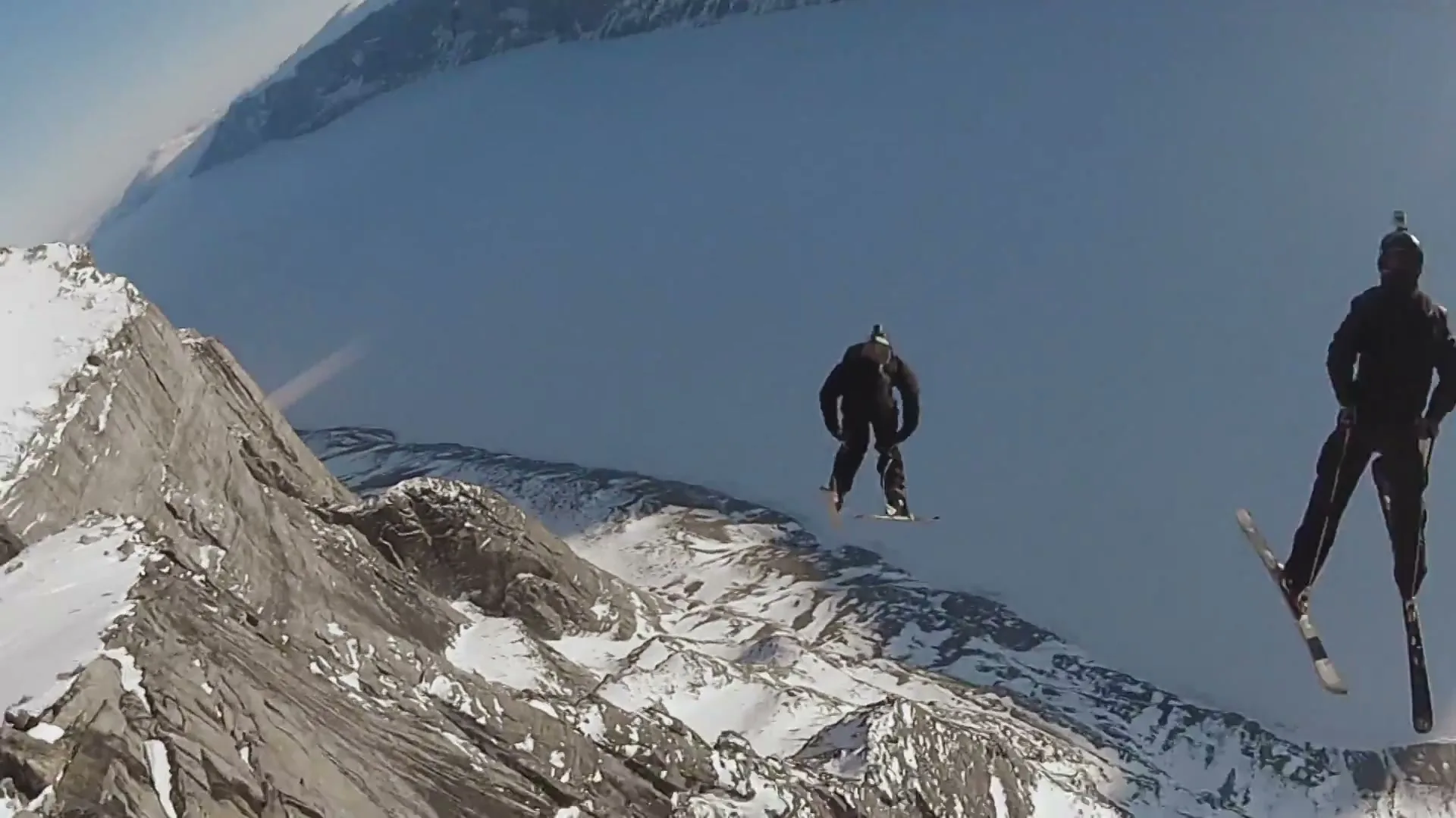 Two skiers in black gear ski off a snowy mountain cliff, falling through the air with a vast frozen lake and mountains in the background.