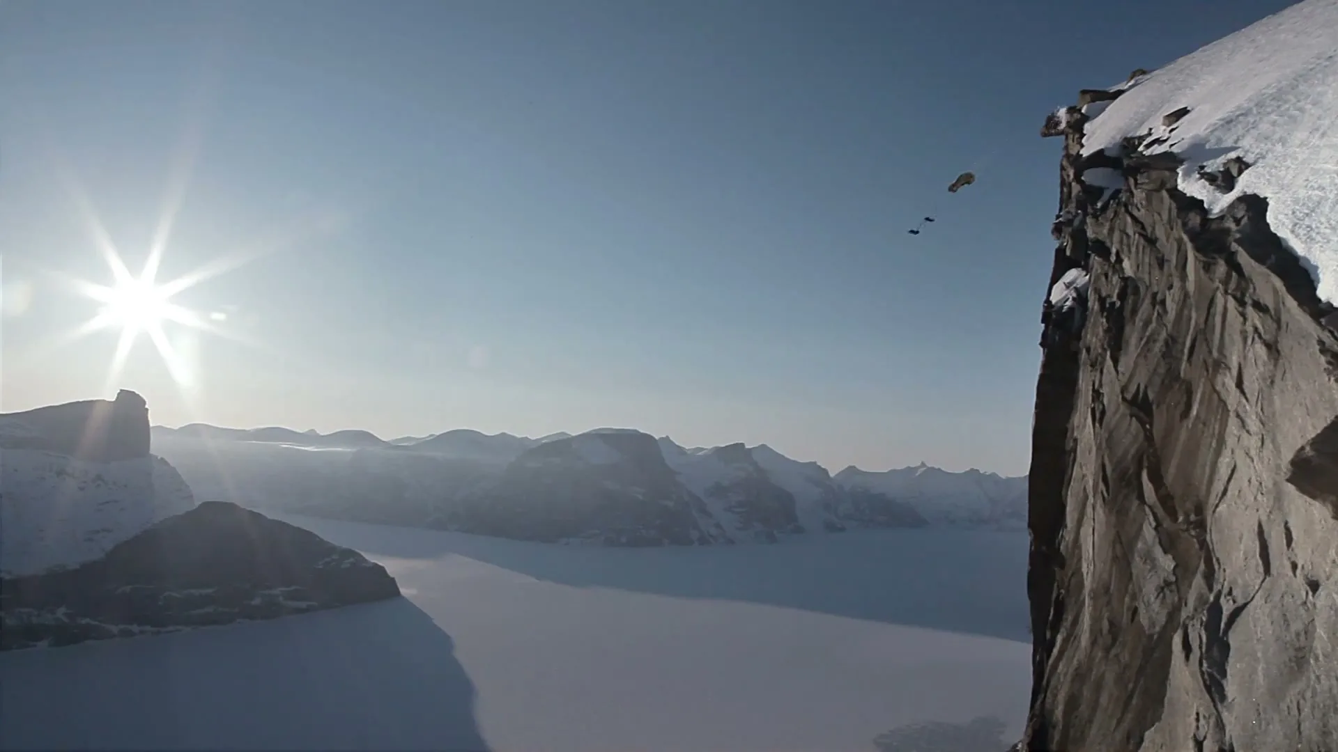 Two base jumpers in black suits leap from a snowy cliff, deploying a yellow parachute against a bright sun and vast, icy landscape.