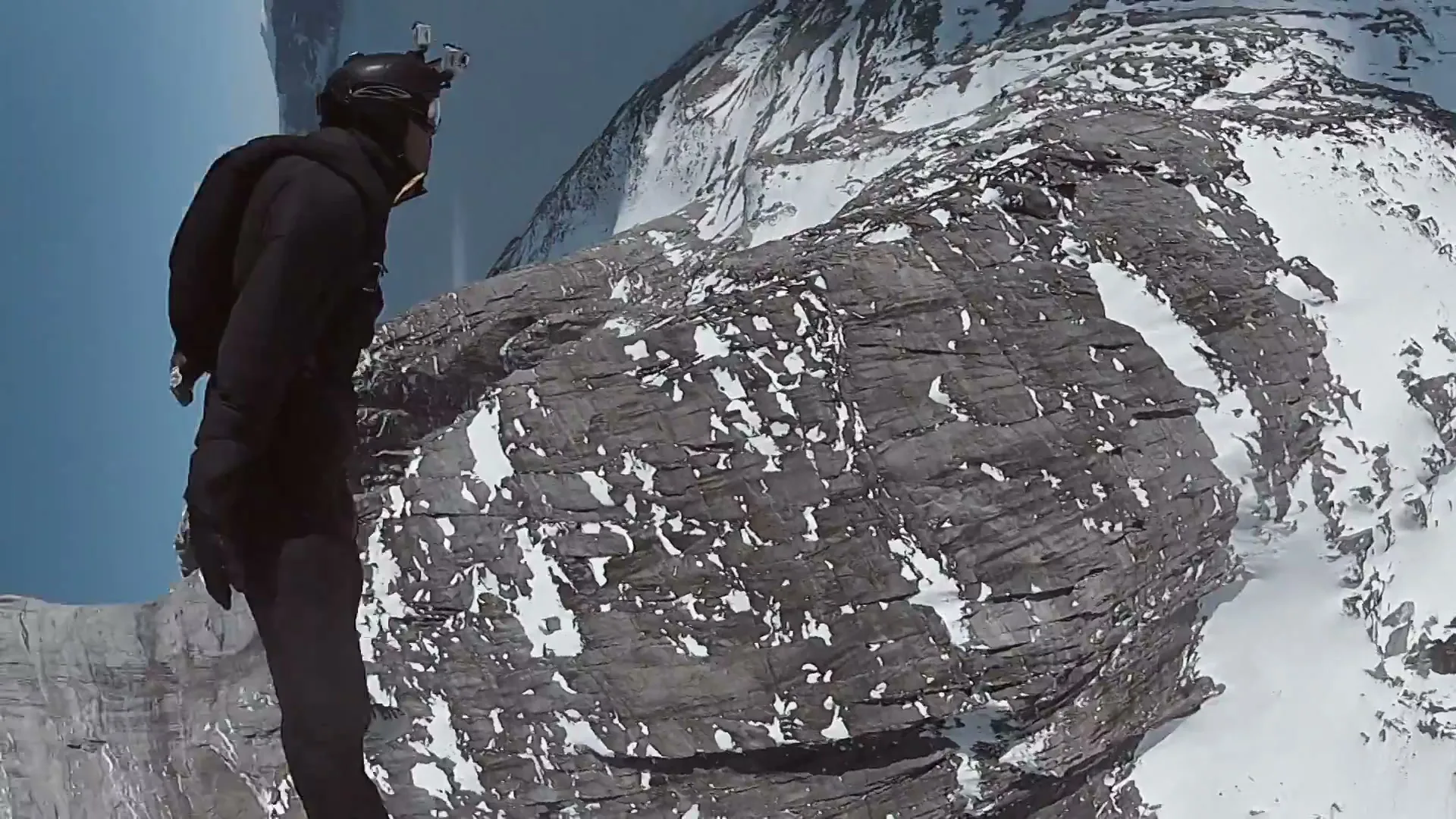 A male base jumper in a black suit and helmet stands on a snowy mountain cliff, then jumps off into the clear blue sky.